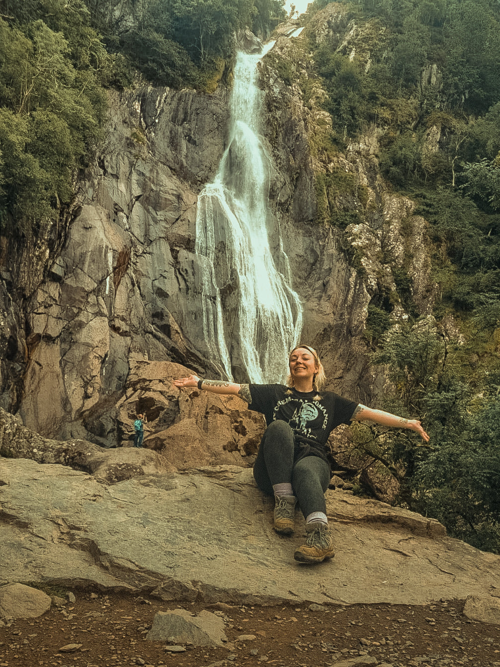 a woman sat smiling with her arms spread with a waterfall in the background